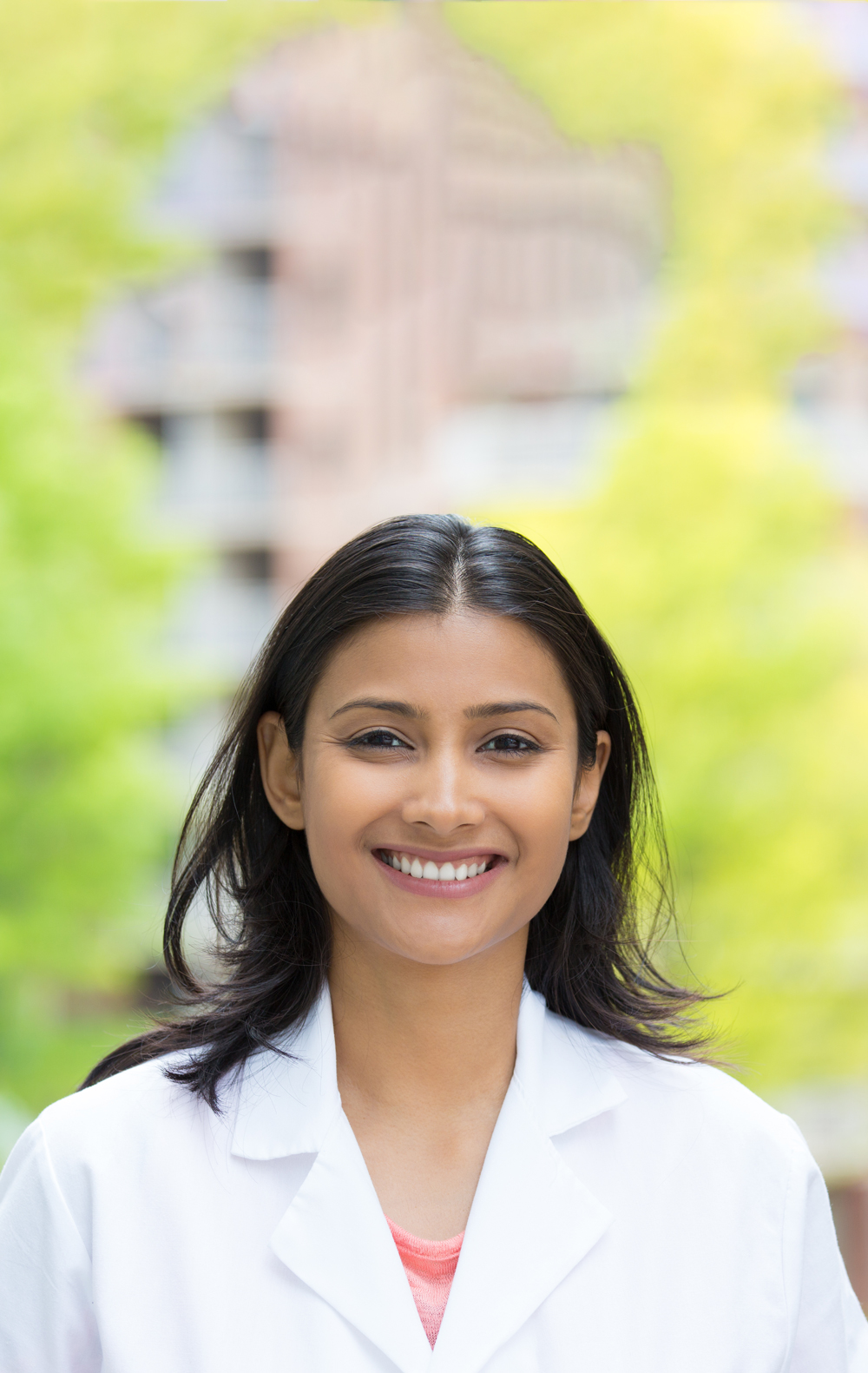 Closeup portrait of confident, smiling female health care professional in white lab coat, isolated background of blurred trees and buildings