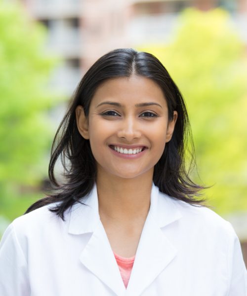 Closeup portrait of confident, smiling female health care professional in white lab coat, isolated background of blurred trees and buildings